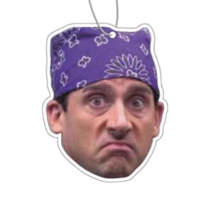 May include: Air freshener featuring a surprised face wearing a purple bandana with a floral pattern. The air freshener has a white border and a silver string for hanging. The face has a concerned expression.