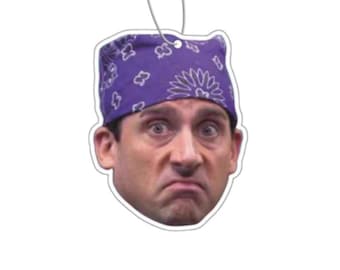 May include: Air freshener featuring a surprised face wearing a purple bandana with a floral pattern. The air freshener has a white border and a silver string for hanging. The face has a concerned expression.