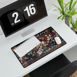 May include: A floral mousepad with a white keyboard and mouse on a white desk. The mousepad features a dark brown background with colorful flowers and green leaves. A black computer monitor displays the time as 2:16 PM.