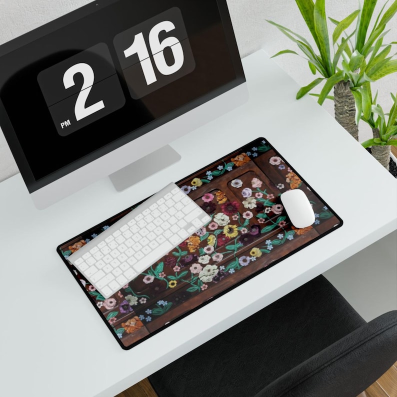 May include: A floral mousepad with a white keyboard and mouse on a white desk. The mousepad features a dark brown background with colorful flowers and green leaves. A black computer monitor displays the time as 2:16 PM.