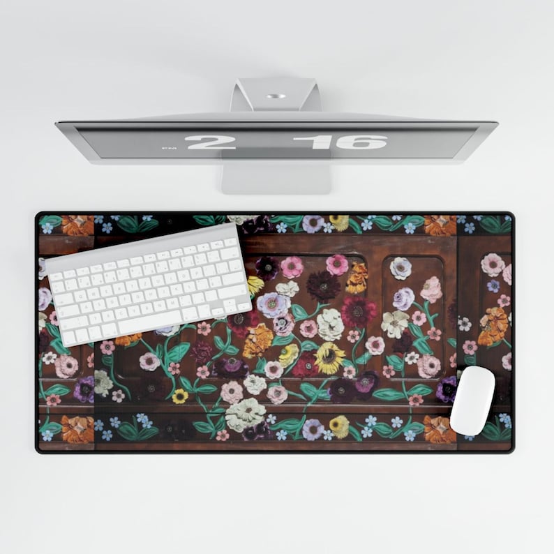 May include: A rectangular desk mat with a floral design featuring a variety of colorful flowers on a dark brown background. A white keyboard and mouse are on the mat, with a computer monitor above.