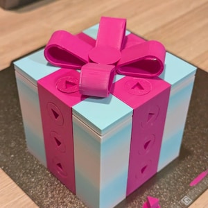 May include: A 3D-printed gift box in light blue with a bright pink ribbon and accents. The box features a bow and vertical pink bands with arrow-shaped cutouts. A small pink triangle is placed beside the box.