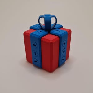 May include: A red, cube-shaped gift box with blue ribbon detailing and a blue handle. The box has a modern, geometric design, with the blue ribbon wrapping around the sides. The box is a solid colour, with no visible text or patterns.