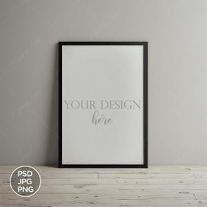 Könnte beinhalten: Ein schwarzer gerahmter Poster-Mockup mit dem Text "YOUR DESIGN here" auf weißem Hintergrund. Der Rahmen lehnt an einer grauen Wand auf einem hellen Holzboden. Das Bild enthält ein PSD-, JPG- und PNG-Logo.
