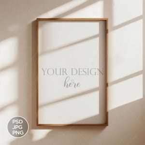 Op de afbeelding: Een lichtbruin houten frame met een witte achtergrond. De tekst "YOUR DESIGN here" is in grijs gedrukt op de witte achtergrond. Het frame bevindt zich op een lichtbeige muur met schaduwen van een raam.