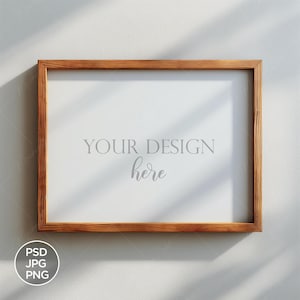 May include: A wooden frame with a white background and the text "YOUR DESIGN here" in gray. The frame is hanging on a white wall with light shining through a window.