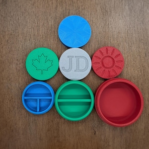 May include: A set of seven round, colourful, plastic containers with various designs. The containers have a variety of designs, including a maple leaf, the letters "JD", a sun, and two containers with dividers.