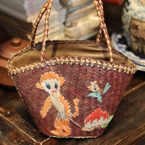May include: A woven brown and beige basket bag with a brown leather handle. The bag has a whimsical design featuring a monkey, a bird, and a flower, all made of colorful woven straw.
