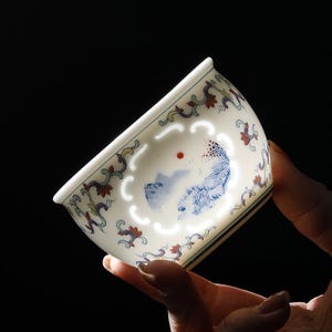 Puede incluir: Taza de té de porcelana blanca con un diseño de paisaje en azul y rojo. La taza tiene una forma curvada y está decorada con motivos florales en el borde. El interior presenta una escena de montaña con un punto rojo.