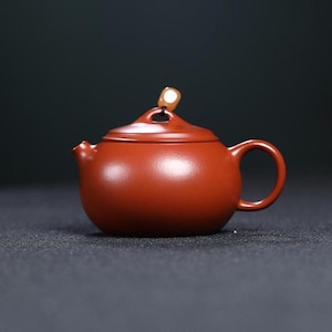 May include: A small, round, reddish-brown ceramic teapot with a lid and handle. The lid has a small, light-colored decorative element. The teapot is designed for brewing tea and has a spout for pouring.