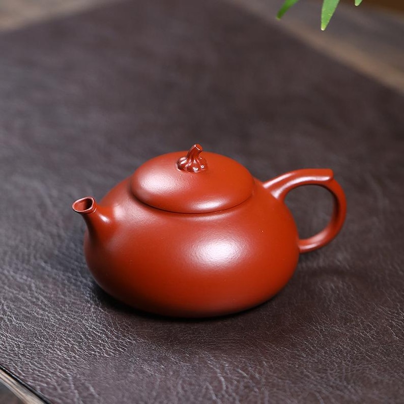 May include: A small, round, reddish-brown teapot with a matching lid and handle. The lid is topped with a decorative knob. The teapot is made of ceramic and has a smooth, matte finish. The spout is small and curved.