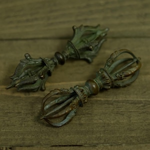 May include: Two antique bronze vajra scepters, featuring detailed floral and geometric patterns. The scepters have a weathered, aged look with a green patina, indicating they are vintage or antique. The background is a wooden surface.