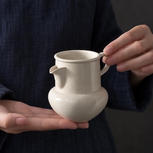 Puede incluir: Una pequeña jarra de cerámica color blanco hueso con una base redondeada y abombada, acabado liso, un pico vertedor corto y un asa lateral. La jarra presenta una sección superior cilíndrica y un cuerpo inferior más ancho y curvo.