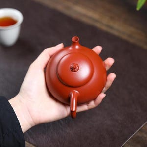 May include: A small, round, reddish-brown teapot with a lid and handle. The teapot is held in a person's hand. A white teacup with tea is in the background. The teapot has a small spout and a decorative element on the lid.