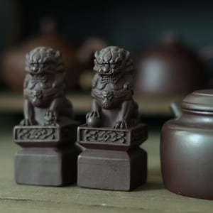 May include: Two dark brown clay guardian lion figurines, each perched atop a square pedestal with intricate carved detailing. The lions feature stylized manes and fierce expressions, with one resting a paw on a sphere and the other on a small cub.