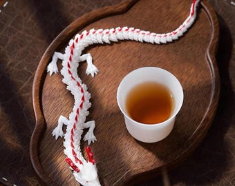 May include: A white and red dragon figurine rests on a wooden tray next to a white teacup filled with tea. The dragon has a detailed design with red accents along its spine and head. The tray has a dark brown finish.