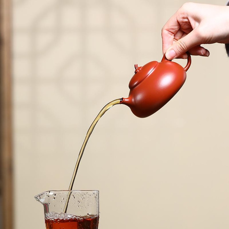 May include: A small, reddish-brown ceramic teapot pouring a stream of tea into a clear glass. The teapot has a rounded body and a curved spout. The tea is a rich amber color, and the glass is partially filled.