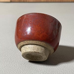 Può includere: Una piccola tazza rotonda in ceramica rossa con una superficie strutturata e smaltata. La tazza ha una base chiara non smaltata. La tazza è probabilmente per bere tè o sake.
