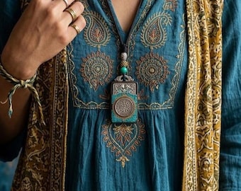 Könnte beinhalten: Türkisfarbene Tunika mit aufwendiger Stickerei und langer Anhängerkette. Die Kette hat einen rechteckigen Anhänger mit türkisfarbenen Akzenten und einem silberfarbenen Design. Eine gemusterte Weste und mehrere Ringe vervollständigen den Boho-Look.