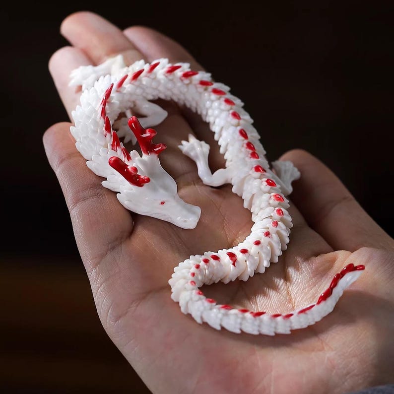 May include: A white dragon figurine with red accents, held in a hand. The dragon has a detailed, segmented body and red markings along its spine and head. The figurine is a decorative item.