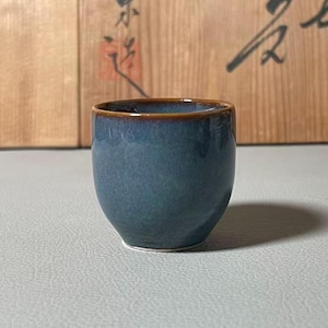 Könnte beinhalten: Eine kleine, blaue Keramiktasse mit abgerundeter Form und braunem Rand. Die Tasse hat eine glänzende Oberfläche und steht auf einer hellgrauen Oberfläche. Der Hintergrund zeigt eine Holzoberfläche mit japanischen Schriftzeichen.