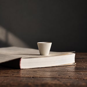 May include: A small, off-white ceramic cup with a tapered, conical shape sits centered on top of a closed, thick book. The cup has a smooth, matte finish and a simple, clean design, resting on the light-colored cover of the book.