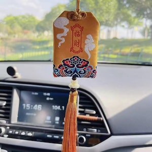 Puede incluir: Un amuleto de coche dorado con un diseño decorativo y una larga borla naranja. El amuleto tiene caracteres chinos y patrones de nubes. Cuelga de una cuerda y está suspendido frente al salpicadero de un coche.