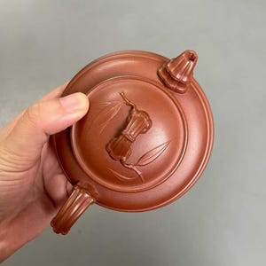 May include: A round, reddish-brown clay teapot lid featuring a central bamboo-shaped handle and embossed bamboo leaf motifs. The lid has a raised circular rim and a small, fluted spout-like protrusion on one side, showcasing a matte texture.
