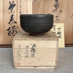 Puede incluir: Un cuenco de té de cerámica negra con forma redondeada, apoyado sobre una caja de madera. La caja tiene caligrafía japonesa y se coloca frente a dos paneles de madera con más caligrafía. El cuenco tiene un acabado oscuro y mate.