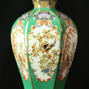 May include: A decorative ceramic vase with a green, white, and gold design. The vase features a detailed pattern of birds, flowers, and trees. The vase has a hexagonal shape and is approximately 12 inches tall.