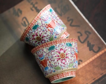 Pode incluir: Duas chávenas de chá decorativas em cerâmica com padrões florais. As chávenas apresentam uma base creme com um design floral vermelho, rosa, azul e verde. As bordas são acentuadas com um padrão geométrico vermelho e dourado. As chávenas estão colocadas numa superfície castanha.