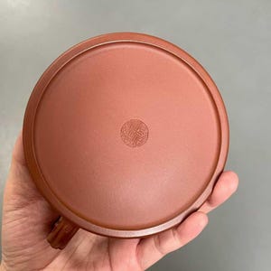 May include: A circular, reddish-brown clay lid for a teapot, featuring a smooth, matte surface and a raised outer rim. In the center of the lid, there is a small, circular stamped seal containing intricate, embossed characters in a traditional style.