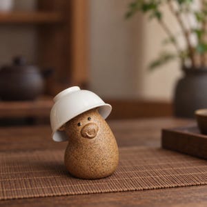 May include: A brown, speckled ceramic tea pet shaped like a bear with a rounded body and a small, smiling face. A smooth, white ceramic bowl is placed upside down on top of the bear's head, covering its ears and resting at a slight angle.