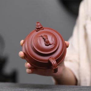 May include: A round, reddish-brown clay teapot featuring a lid with a sculpted bamboo-style handle and embossed bamboo leaf patterns. The teapot has a matching curved handle and a small spout, showcasing a smooth, matte finish throughout the body.