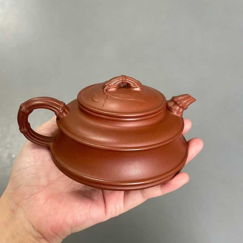 May include: A reddish-brown clay teapot featuring a low, wide body, a lid with a sculpted bamboo-style handle, and a matching bamboo-textured side handle. The spout is short and tapered, and the lid is decorated with a subtle leaf engraving.