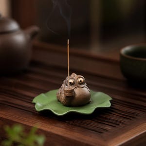 Puede incluir: Un caprichoso porta incienso de rana hecho de cerámica marrón, apoyado en un plato en forma de hoja verde. La rana tiene ojos grandes y expresivos y una pipa. Un palito de incienso encendido emite una voluta de humo. El soporte está sobre una bandeja de madera.