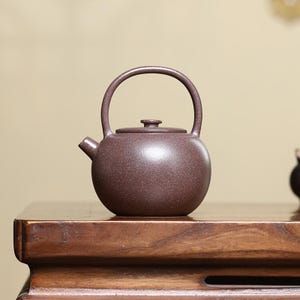 May include: A round, dark brown teapot with a textured, matte finish. The teapot features a single high-arched handle extending from the top, a small spout on the left side, and a circular lid with a small knob centered on the top of the vessel.