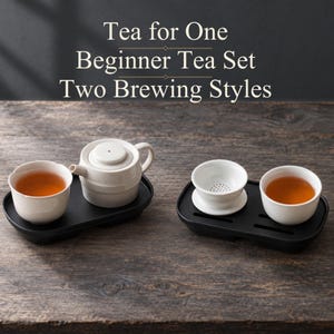 Puede incluir: Un juego de té para una persona con dos estilos de preparación, con piezas de cerámica blanca sobre bandejas negras. Incluye una tetera, dos tazas blancas con té ámbar y un colador. El texto dice: Tea for One Beginner Tea Set Two Brewing Styles.