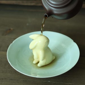 May include: A dark brown teapot pours dark liquid onto a cream-colored ceramic rabbit figurine centered on a pale green saucer. The rabbit is sculpted in a seated position with long ears, and the liquid flows over its head into the shallow dish.
