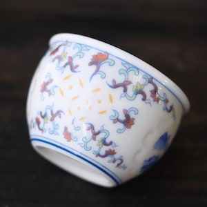 Może przedstawiać: Biała ceramiczna miska z niebieską obw&oacute;dką i skomplikowanymi wzorami kwiatowymi w kolorze pomarańczowym, niebieskim i ż&oacute;łtym. Miska ma zaokrąglony kształt i delikatny design, idealna do serwowania małych porcji.