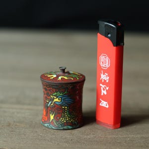 May include: A small, ornate, red and bronze container with a lid, featuring a colorful dragon design. Next to it is a bright red lighter with white text. The items are on a wooden surface.