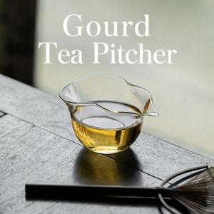 Op de afbeelding: Een heldere glazen theekan met een kalebasvormig lichaam en een kleine schenktuit, gevuld met een goudkleurige vloeistof. De tekst Gourd Tea Pitcher staat in witte letters boven het vat naast een donker handvat van een garde.