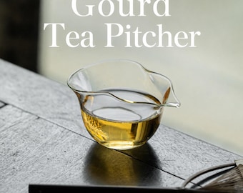 Może przedstawiać: Dzbanek do herbaty z przezroczystego szkła o korpusie w kształcie tykwy z małym dzióbkiem, zawierający złoty płyn. Tekst Gourd Tea Pitcher jest wyświetlany białymi literami nad naczyniem obok ciemnego uchwytu trzepaczki.