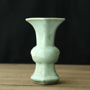 May include: A celadon-colored ceramic vase with a flared top and a bulbous midsection. The vase has a smooth, glossy finish and a slightly irregular shape. The vase is displayed on a wooden surface against a black background.