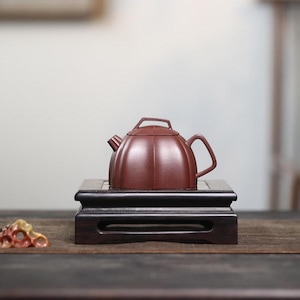 May include: A small, maroon-colored teapot with a curved handle and a lid, resting on a dark wooden stand. The teapot has a rounded body with vertical ridges. The stand is square with decorative edges, and the background is blurred.