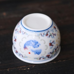 Może przedstawiać: Biała ceramiczna filiżanka do herbaty o zaokrąglonym kształcie, z niebieskimi i czerwonymi wzorami kwiatowymi i centralnym niebieskim wzorem. Filiżanka ma niebieską obw&oacute;dkę. Ustawiona na ciemnej drewnianej powierzchni.