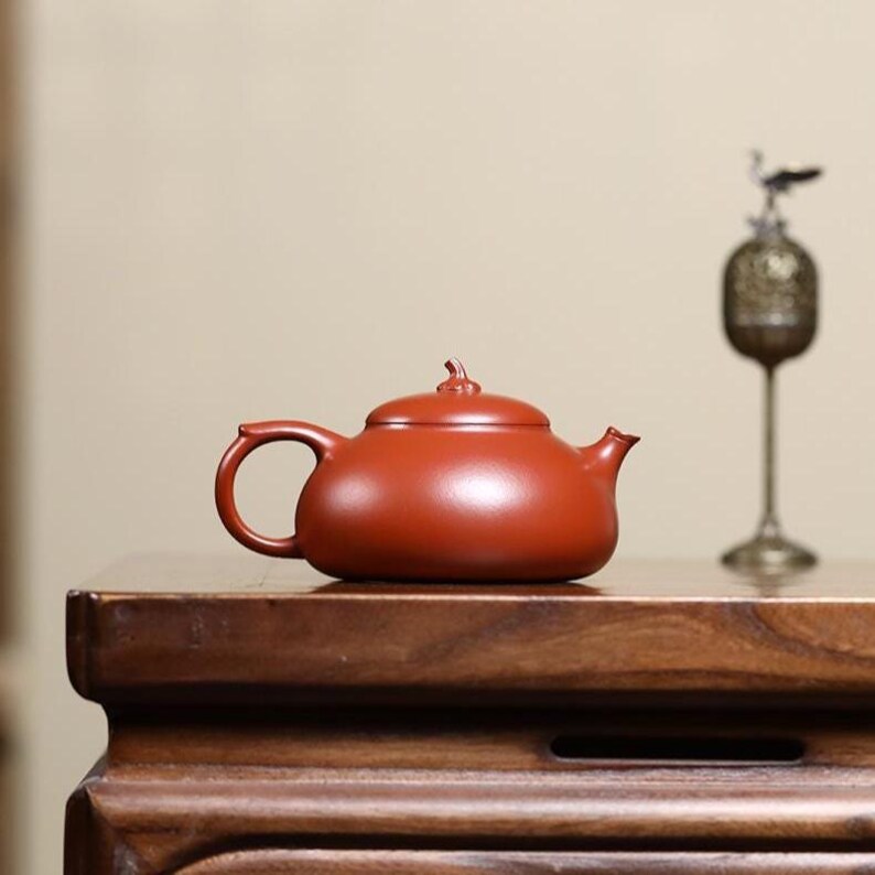 May include: A small, reddish-brown ceramic teapot with a rounded body, a curved handle, and a small spout. The teapot sits on a dark wooden surface. A decorative metal object is in the background.