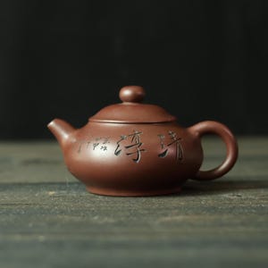 May include: A small, brown ceramic teapot with a rounded body, a curved spout, and a loop handle. The lid has a small knob. The teapot has engraved Chinese characters on the side. The teapot is sitting on a dark wooden surface.