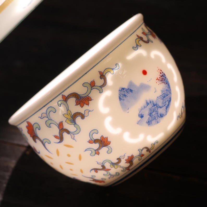 Może przedstawiać: Kremowa ceramiczna miska ze szczeg&oacute;łowym wzorem. Miska ma niebieski i pomarańczowy wz&oacute;r kwiatowy na obrzeżu oraz malowniczą ilustrację g&oacute;r i budynk&oacute;w. Miska ma białą obw&oacute;dkę i ciemne tło.