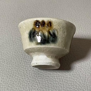 Könnte beinhalten: Eine kleine, cremefarbene Sake-Tasse aus Keramik mit ausgestelltem Rand und Fuß. Die Tasse zeigt ein handgemaltes Design von drei Figuren in Braun- und Schwarztönen. Die Oberfläche hat eine Craquelé-Glasur.
